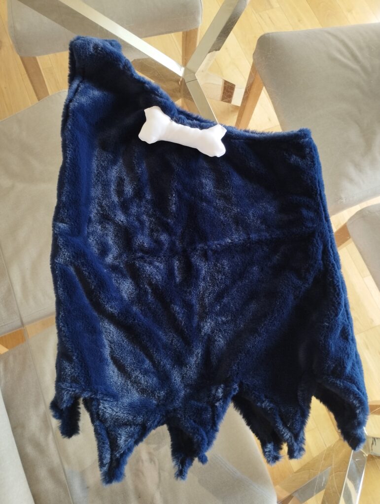 Disfraz de troglodita casero terminado, hecho con tela tipo peluche azul y un adorno en forma de hueso blanco. Diseño rústico con bordes irregulares, ideal para disfraces infantiles de cavernícola o prehistórico.