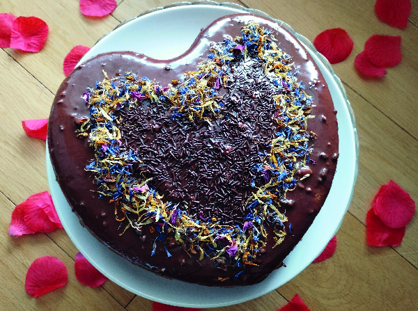 Tarta de chocolate finalizada, decorada con pétalos de flores secas y fideos de chocolate en el centro, presentada sobre un plato blanco con pétalos de rosa alrededor.