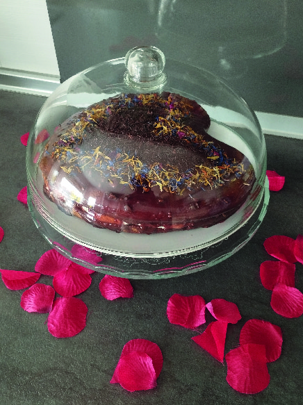 Tarta de San Valentín con cobertura de chocolate y decoraciones dentro de una campana de cristal sobre una mesa con pétalos de rosa, lista para servir.