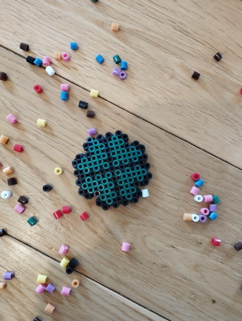 Manualidad de trébol hecho con Hama Beads en tonos verdes y negros, rodeado de cuentas de colores sobre una superficie de madera, ideal para St. Patrick’s Day.
