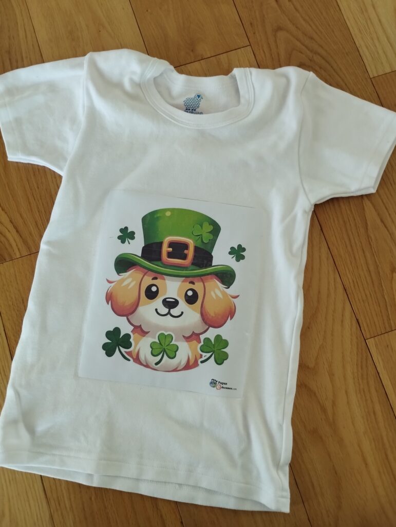 Camiseta blanca con diseño transferido de un perrito con sombrero de duende y tréboles, ideal para manualidades de St. Patrick’s Day.